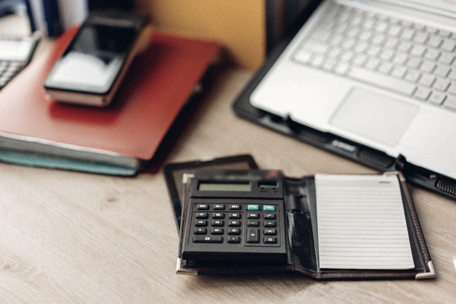 Mesa de trabajo con una calculadora sobre una libreta, un móvil sobre una carpeta granate y un portátil abierto a la derecha.
