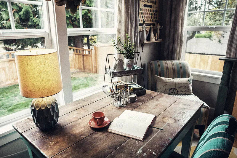 Rincón de trabajo en casa: mesa de madera junto a una ventana, con una lámpara de pantalla ocre, una taza roja y un cuaderno abierto.
