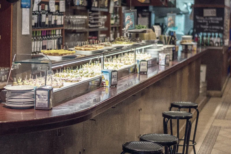 Barra de un bar de tapas de Barcelona, con una vitrina de pinchos, platos apilados y taburetes negros en primer plano.